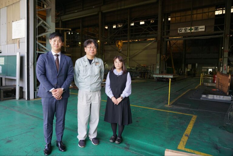 菅原工業の工場現場に立つソフト開発課の越原様、総務・経理部の橋本様と白石担当者